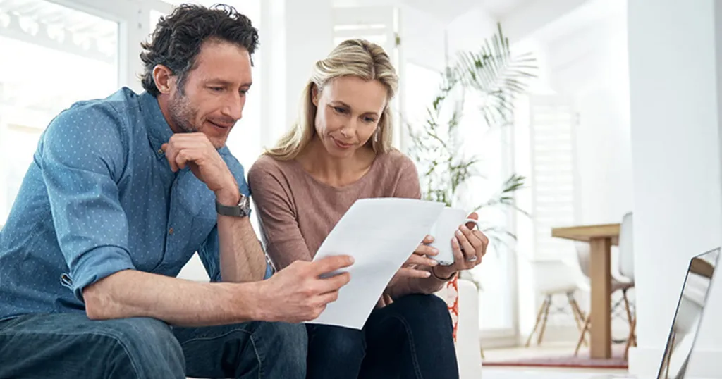A middle-aged couple sitting on a step in their home, looking over documents.
