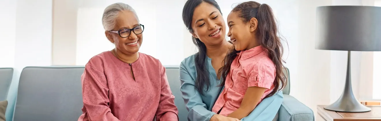 A grandmother, daughter and granddaughter sit and smile together on a couch and consider the Mutual of Omaha provides Life insurance options for all three generations.  