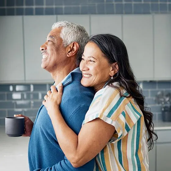 A woman embraces her husband as he enjoys a cup of coffee, and she is happy they are covered with whole life insurance. 