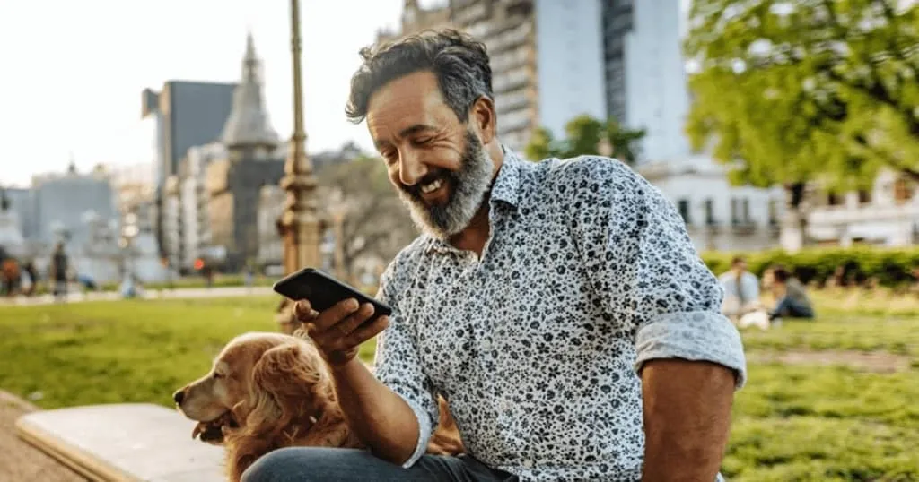 Man sitting in the park with his dog, smiling at his phone. 
