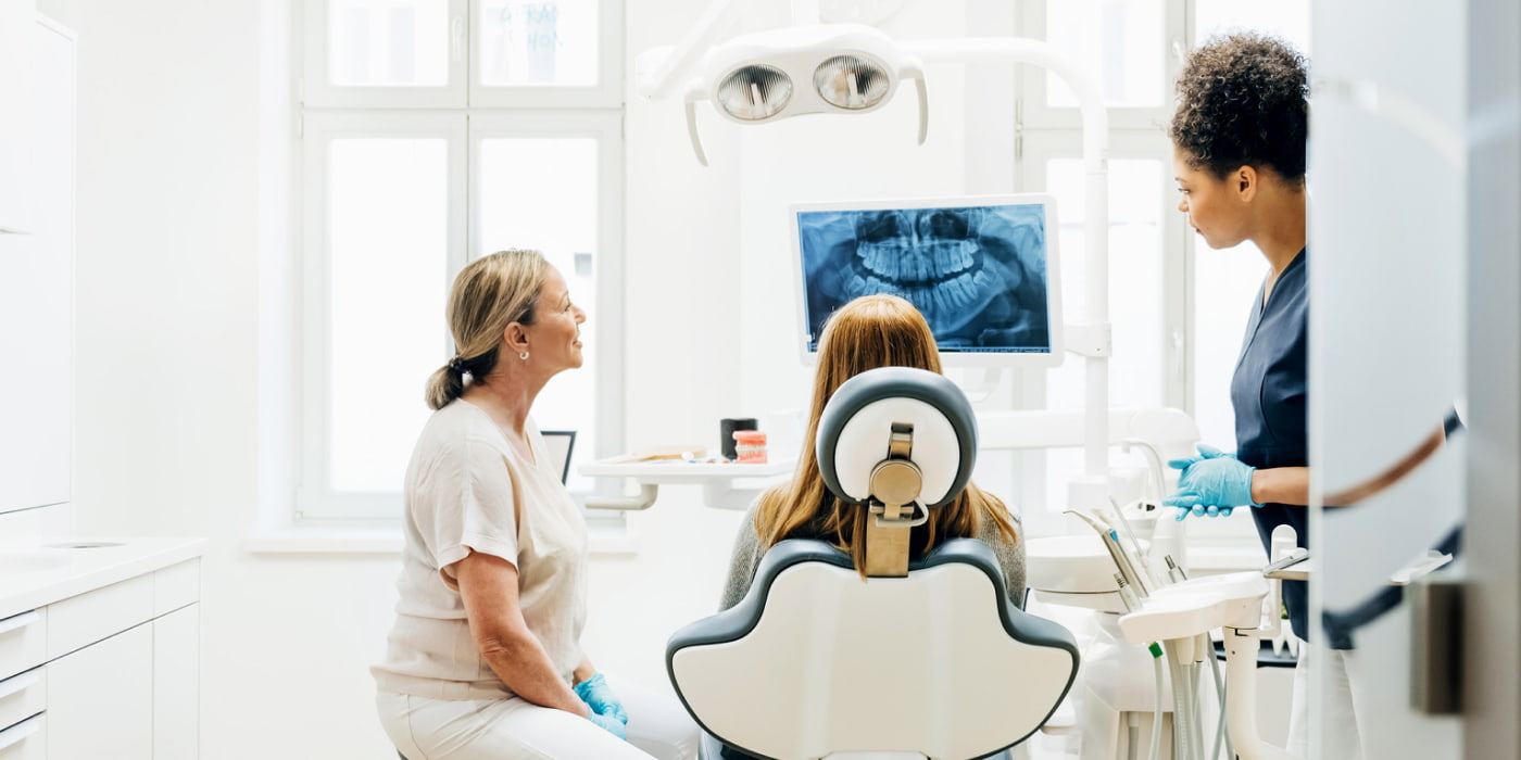 Three women in a bright dental office gather around a dental chair as a patient sits facing a monitor displaying dental X-rays, while two dental professionals stand nearby with tools and gloves, discussing treatment.