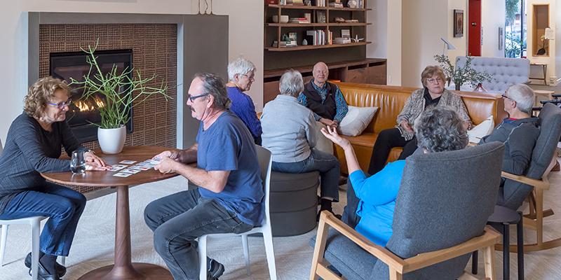 A group of eight seniors sit in small groups in a common area at PDX Commons cohousing community. Two people sit at a table playing cards, while other groups sit and talk together.