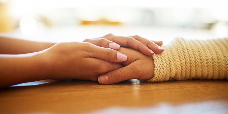 A close-up of a woman’s hands gently holding another woman’s hand as they both rest on a tabletop.