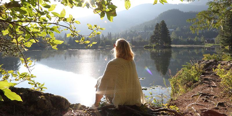 A woman wearing a shawl sits on a the ground looking out over a small, peaceful lake. The sun is shining through tree branches in the foreground and there are tree-covered hills on the other side of the lake.