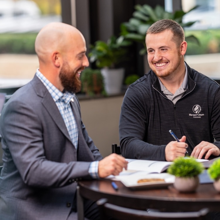 Two professionals sit at a small round table in an office setting, reviewing documents and taking notes during a financial planning discussion, with plants and large windows in the background.