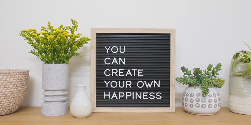 A small decorative sign that says "You can create your own happiness" sits on a table between two plants and a woven basket.