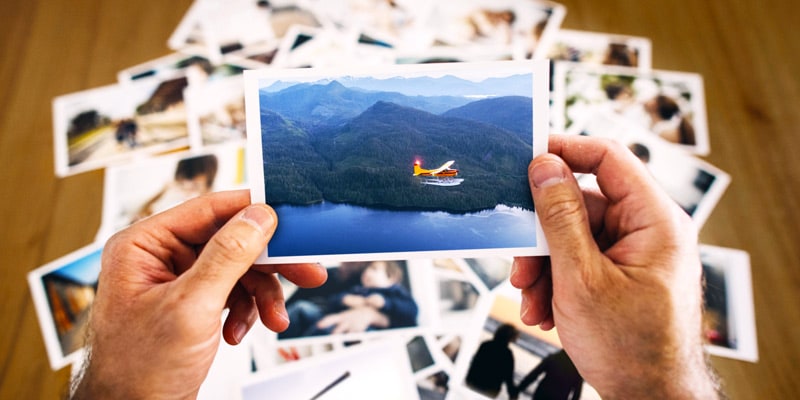 A close-up of a man's hands holding an old color photograph of a red seaplane flying over a lake with mountains in the background. Behind the man's hands is a table with a pile of similar photos on it.