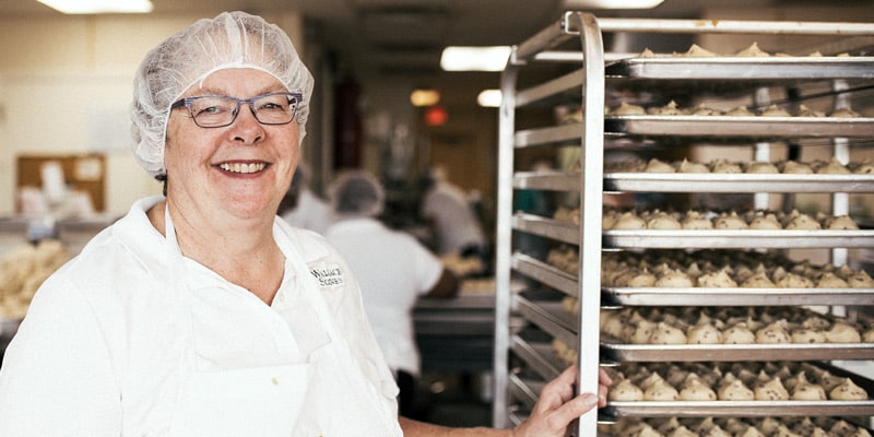 Mary Wallace, of Wallace Scones, dressed in a hair net and apron, stands next to a rack filled with trays of unbaked scones. Other workers are in the background